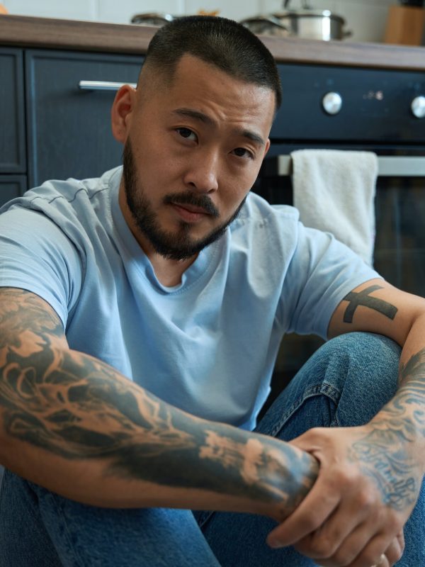 Young asian man sits on floor in kitchen, hugging his knees